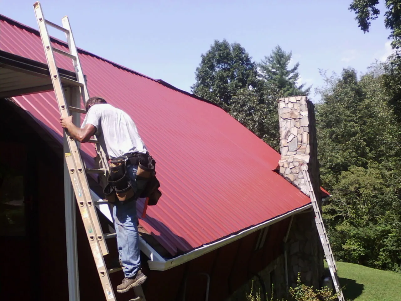 Roofer on a ladder beside a red metal roof and stone chimney — Soffit & Fascia Repair by Roofing Brothers in Deer Island